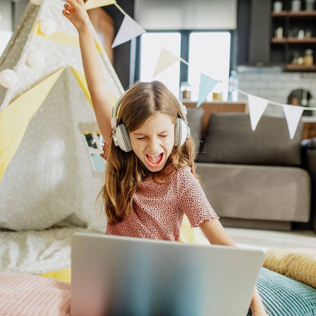 A little cute girl is on the floor in front of her tent in the living room, she enjoys using a laptop and listening to music